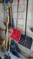 Group of garden tools leaning against wall: rakes, shovels, snow shovels, brooms