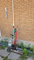 Group of four tools leaning against wall: pole pruner, string trimmer, shovel, push broom