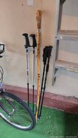 Group of five poles leaning against wall, including wood stick and four metal trekking poles