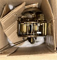 Clock movement in cardboard box showing overall form and attached pendulum leader rod