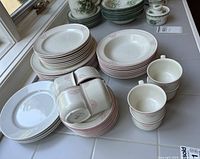 Assorted stacks of pink-rim dinnerware, green floral plates, plain white plates, cups, sugar bowl, creamer