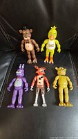 All five FNAF articulated figures laid out on black leather: Freddy, Chica, Bonnie, Foxy, Golden Freddy