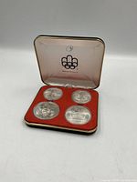 Four coins in red-lined clamshell case, lid open, Olympic logo visible