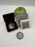 Canadian proof coin in open clamshell case alongside box, certificate, Chinese coin in plastic, green apple for scale