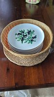 Stack of six fruit-motif plates inside wicker basket on woven mat