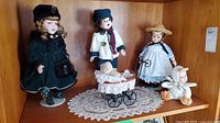 Shelf view showing three standing porcelain dolls, miniature stroller with baby figure, baby doll in duck suit