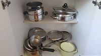 Cabinet shelf showing stainless steamer set, sauté pan with lid, nested saucepans, lids, and yellow enamel pot