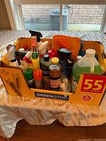 Overall view of box containing assorted cleaning supplies, showing spray bottles, liquid cleaners, aerosol cans, cloths