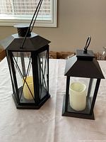 Two black metal lanterns, hexagonal and square, each with a white pillar candle inside, placed on table