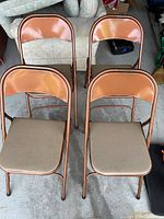 Four copper/bronze metal folding chairs with beige vinyl seats, front view