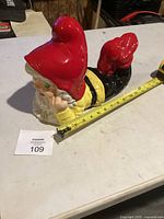 Top view of ceramic gnome showing red hat, yellow shirt, black pants; tape measure indicates about 13 in length