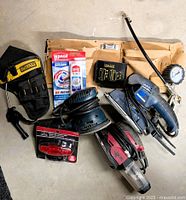 All items arranged on floor showing sanders, tool belt, holster, air tools, hinge plates, caulk tube and bike light