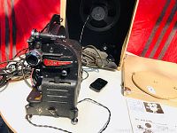 Photo showing the Pathoscope Son projector with a black textured metal body, lens, power cords, placed near the open carry case and instruction booklet.