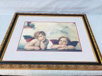 Full view of the framed angel cherub print showing two cherubs resting on a ledge with a cloudy sky background, set in gold and black frame.