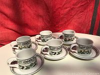 Set of six coffee mugs each paired with a matching underplate, displayed against a red fabric background, showing unique botanical floral designs on each mug and underplate.