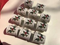 Twelve white coffee mugs decorated with green holly leaves and red berries arranged in rows on a white surface.