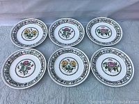 Photo showing the full set of seven vintage Portmeirion Variations dinner plates laid out in two rows, clearly displaying the floral and butterfly designs and the decorative floral vine border around each plate's rim.
