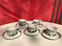 Front view of 5 tea cups each paired with a saucer showing floral and butterfly designs on cream background with pink and green borders.