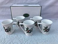 Set of five white ceramic Portmeirion coffee mugs with holly and ivy design arranged on white quilted fabric with original Portmeirion box in background.
