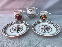 Two floral pattern plates, a cream pitcher with text, and two mugs with fruit illustrations arranged on quilted fabric background.