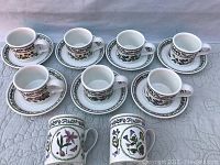Seven porcelain mugs and seven matching saucers laid out on a textured cloth surface, showing the floral designs and leaf borders.