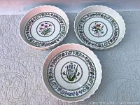 Three white ceramic serving dishes with scalloped edges, each featuring a different floral design and surrounding leaf and flower border pattern, laid out side by side on a quilted surface.