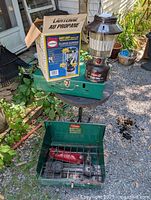 Lot overview showing two green Coleman stoves, brown Coleman Eas-Lite lantern, and boxed Century Primus propane lantern