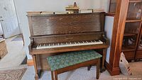 Front view of oak upright piano with green upholstered bench, piano lamp and books on top