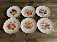 Six fruit motif plates displayed face up , showing designs and gold scalloped rims