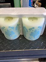 Front view of two pale green-ivory wax LED candles with blue damask design in styrofoam tray