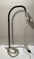Full view of silver floor magnifying lamp showing base, gooseneck arm, magnifier head and power cord