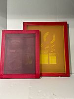 Front view of two red wooden silkscreen frames showing mesh colors and printed designs