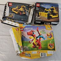 Three LEGO boxes: damaged Technic box back view, intact Technic Bulldozer 42163 front, damaged Creator dragon 3-in-1 front
