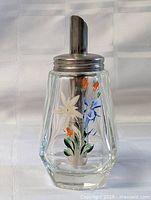 Full view of glass sugar shaker showing faceted shape, floral decoration, metal lid and spout