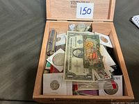 Open cigar box displaying contents: coins, banknotes, stamps