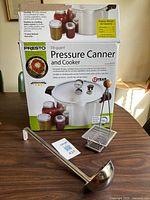 Front view of boxed Presto pressure canner with ladle and jar lifter displayed