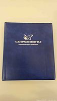 Front of blue binder with title and shuttle logo