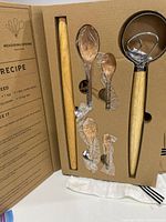 Open box showing rolling pin, whisk, four wooden measuring spoons still wrapped