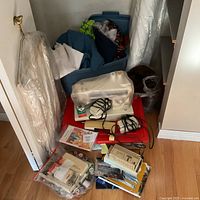 Closet area with garment bag, blue bin of fabric, red tote lid holding sewing machine and accessories