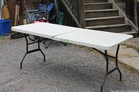 Full view of white 8-ft folding table set up on gravel surface showing center hinge and black legs