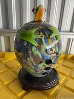 Front angled view of multicolored glass vase with green frog lid on wooden base