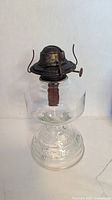 Full view of clear glass oil lamp showing embossed base, fuel font, metal burner with wick, no chimney