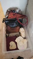 Clear bin containing tapestry handbag, black handbag, brown bag, burgundy dog motif bag, scrunchie, and baseball cap