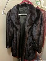 Front view of dark brown/black Pappas fur coat on hanger with black leather gloves tucked in and dust cover visible behind