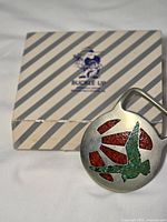 Front view of round pewter buckle with turquoise and red stone inlay, Buckle Up box behind