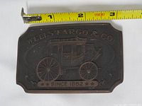 Front of brass Wells Fargo belt buckle showing stagecoach design and tape measure indicating 3.5 in width