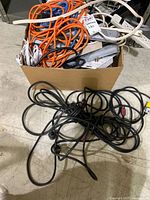Box of assorted orange, blue, clear extension cords with power strips; black cord bundle on floor