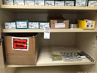 Wide view of cabinet shelf showing multiple Spaenaur boxes, cardboard carton, plastic tray with aluminum spacers and bags of washers