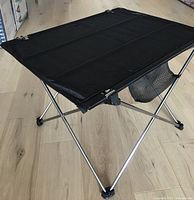 Angled view showing black fabric tabletop, silver X-frame legs, mesh pocket under table