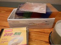 Box of Memorex slim jewel cases with multi-color cases, Staples DVD-RW package, portion of CD-R spindle